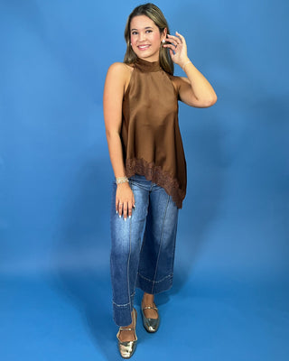 A woman modeling a sleeveless high-neck brown satin top with a lace-trimmed hem, paired with blue jeans and metallic gold shoes, against a solid blue studio backdrop.