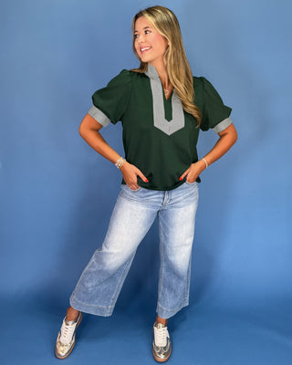 Woman wearing a green top with gray accents and blue jeans against a blue background
