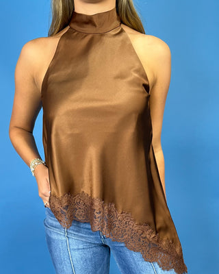 A woman wearing a brown satin halter top with a lace hem, paired with blue jeans, set against a solid blue background.