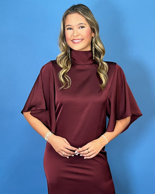 Burgundy satin midi dress with a turtleneck, flutter sleeves, relaxed silhouette, and side slit detail.