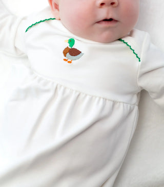 Baby wearing a white outfit with a embroidered duck design on a plain background