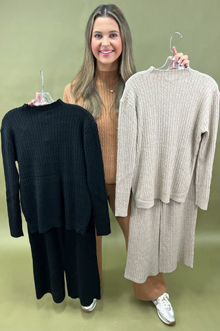 Woman holding two sweaters, one black and one beige, on hangers against a green background.
