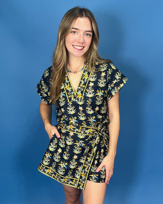 wearing a short black wrap dress with a yellow and white botanical pattern.