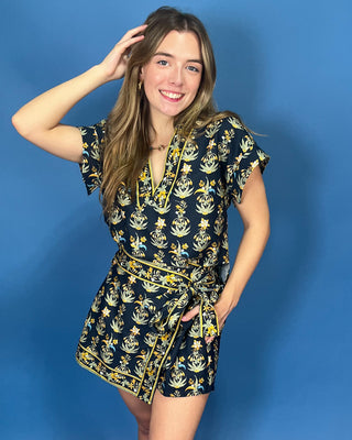 A woman modeling a short-sleeve wrap romper with a navy blue base and a yellow and white botanical pattern