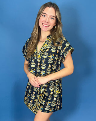 A smiling young woman with long light brown hair stands wearing a short navy wrap dress with a yellow botanical pattern and yellow piping.