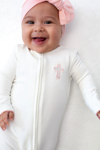 Baby wearing a white onesie with a pink cross design and a matching pink headband.