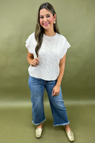 Woman wearing a white cable knit top and blue jeans on a green background
