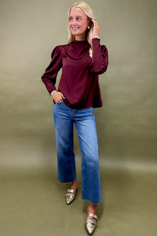 Woman wearing a burgundy blouse and blue jeans against a green background