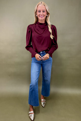 Woman wearing a burgundy blouse and blue jeans against a plain background
