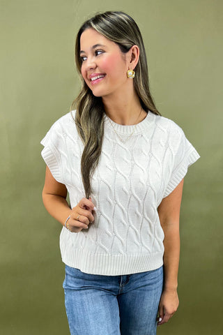 Woman wearing a white cable knit sweater and blue jeans against a green background