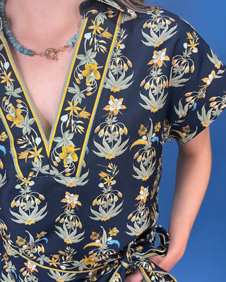 A close-up of a person wearing a navy wrap-style blouse with a yellow, white, and sage green floral print, paired with a blue beaded necklace