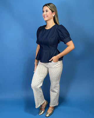 A woman modeling a navy blue short-sleeve peplum top and light gray textured wide-leg pants against a solid blue studio backdrop.
