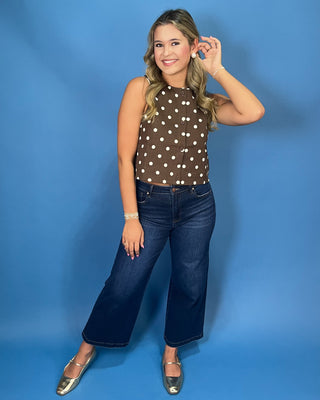 A woman modeling a brown sleeveless top with white polka dots, paired with dark blue wide-leg jeans and metallic silver flats