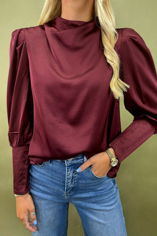 Person wearing a burgundy blouse with puffed sleeves and blue jeans against a green background