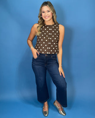 A woman modeling a brown sleeveless top with white polka dots, paired with dark blue jeans and metallic silver flats