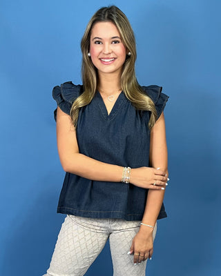 A smiling young woman with blonde-highlighted hair stands against a solid blue studio background, wearing a dark navy ruffled sleeveless top and light gray quilted pants.
