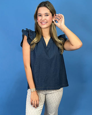 A woman modeling a sleeveless dark blue denim top with ruffled shoulders and a V-neckline, paired with light gray patterned pants, set against a solid blue studio background.