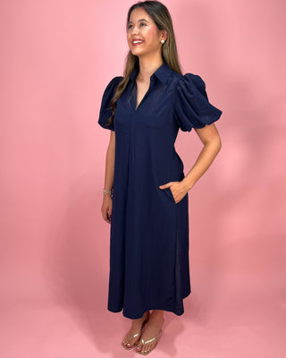 Woman wearing a navy dress with puff sleeves against a pink background