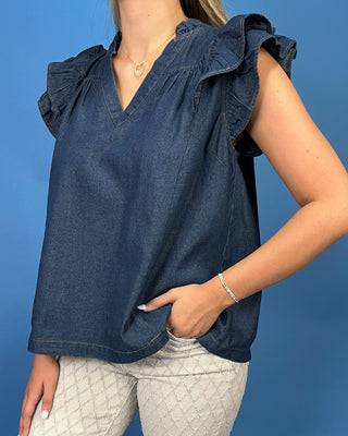 A woman wearing a dark blue denim ruffle-sleeve top and light gray patterned pants, standing against a solid blue background.