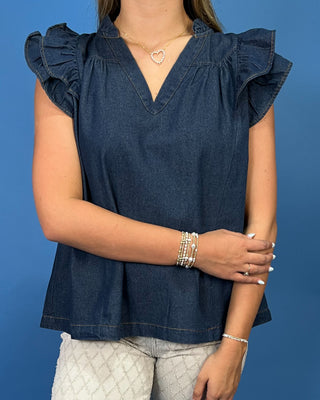 A woman wearing a dark blue denim top with ruffled cap sleeves and a V-neckline, paired with light gray patterned pants, set against a solid blue background.