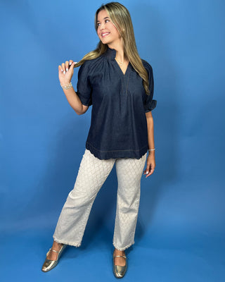 A woman modeling a dark navy blue short-sleeve top with a gathered neckline and V-neckline, paired with light beige patterned pants and metallic gold flats, set against a solid blue studio background.