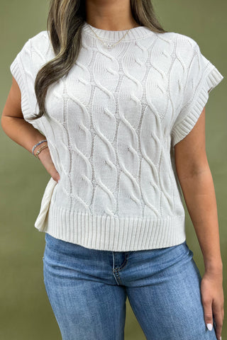 Woman wearing a white cable knit sleeveless sweater and blue jeans against a green background