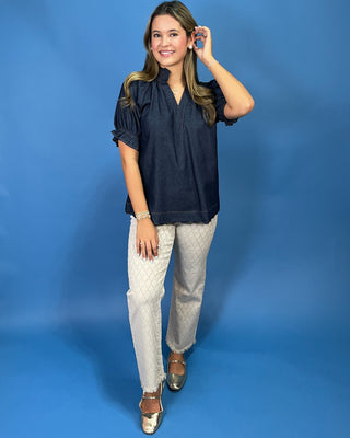 A woman modeling a dark navy blue short-sleeve blouse with puff sleeves and a gathered neckline, paired with light beige patterned pants and metallic shoes, set against a solid blue studio backdrop.