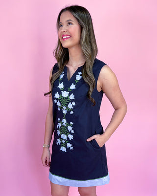 Woman wearing a navy sleeveless dress with floral embroidery on a pink background