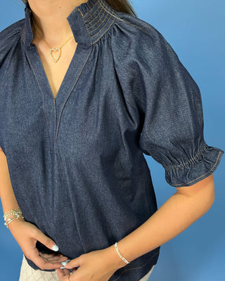 A woman wearing a dark blue denim short-sleeve blouse with a smocked collar and gathered sleeves, paired with a gold heart necklace and bracelets, set against a solid blue background.