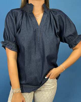 A woman wearing a dark blue chambray puff-sleeve blouse with a gathered mock neck and smocked cuffs, paired with light-colored patterned pants, set against a solid blue background.