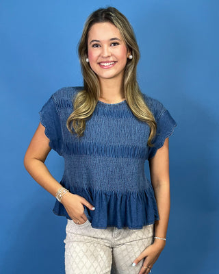A woman modeling a blue smocked short-sleeve top with a ruffled hem, paired with light beige patterned pants, standing against a solid blue studio background.
