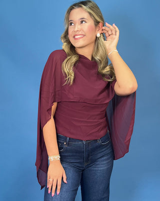 A woman modeling a draped, short-sleeved top in a deep burgundy or maroon color, paired with blue jeans