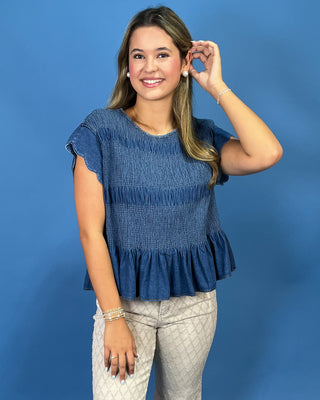 A woman modeling a blue smocked short-sleeve peplum top with light beige patterned pants, standing against a solid blue background.