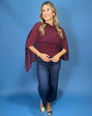 A woman modeling a burgundy draped cape top with wide short sleeves, paired with dark blue jeans and metallic gold flats
