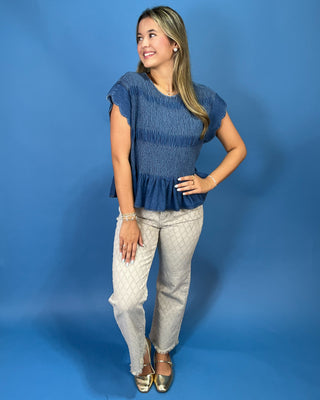 A woman posing in a blue textured short-sleeve peplum top and light beige patterned trousers against a solid blue studio backdrop.