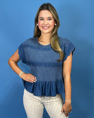 A woman modeling a blue smocked short-sleeve top with a ruffled hem, paired with light beige patterned pants, against a solid blue studio background.
