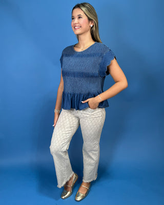 A woman modeling a blue textured short-sleeve top with a ruffled hem and light gray patterned pants, standing against a solid blue studio backdrop.
