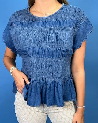 A woman wearing a blue smocked short-sleeve top with a ruffled hem, paired with light gray patterned pants, standing against a solid blue background.