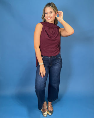 A woman modeling a sleeveless draped top in deep burgundy, paired with dark blue jeans and metallic gold flat shoes