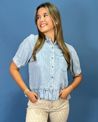 A woman wearing a light blue chambray short-sleeve button-up blouse with a ruffled hem and beige quilted pants, posing against a solid blue studio background.