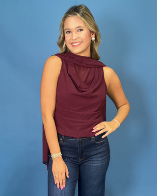 A woman modeling a sleeveless draped top in deep burgundy, paired with dark blue jeans