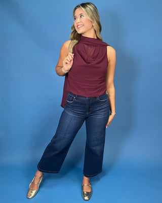A woman modeling a sleeveless draped cowl-neck top in burgundy, paired with dark blue jeans and metallic gold flats
