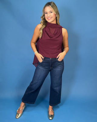 A woman modeling a sleeveless draped top in deep burgundy, paired with dark blue jeans and metallic gold flats