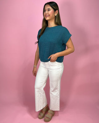 Woman wearing a teal top and white pants against a pink background