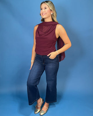 A woman modeling a sleeveless draped top in deep burgundy paired with dark blue jeans and metallic gold flats