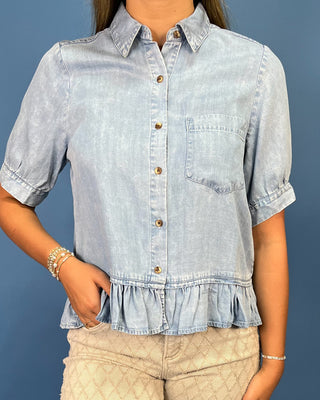 A woman wearing a light blue denim short-sleeve button-up shirt with a ruffled hem and a chest pocket, paired with beige quilted pants, set against a solid blue background.