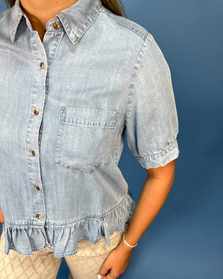 A person wearing a light blue chambray short-sleeve button-up shirt with a ruffled hem and chest pocket, paired with cream patterned pants, against a solid blue background.