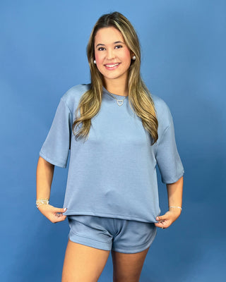 A woman modeling a matching light blue lounge set consisting of a short-sleeve crewneck top and matching shorts, posed against a solid blue studio backdrop.