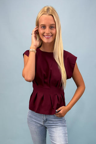 Woman wearing a burgundy peplum top and light blue jeans against a gray background