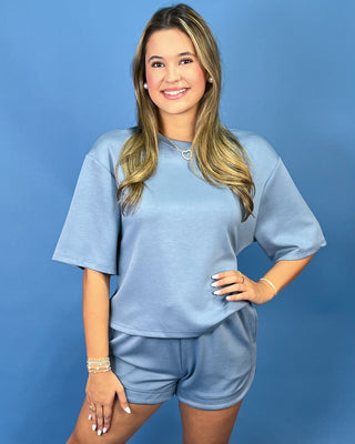 A woman modeling a light blue lounge set consisting of a short-sleeve crewneck top and matching shorts, posed against a solid blue studio background.